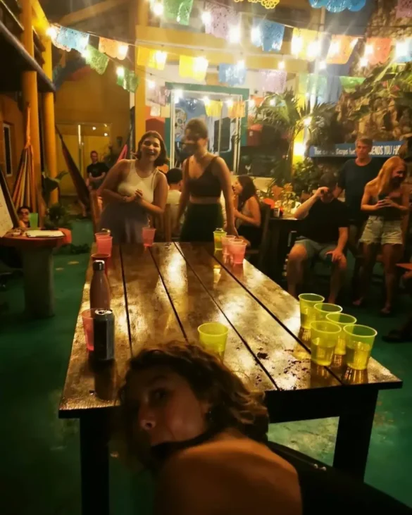 Someone leaning down under a camera while a photo is taken of people playing beer pong in a large courtyard with Mexican flags ahead and people enjoying themselves