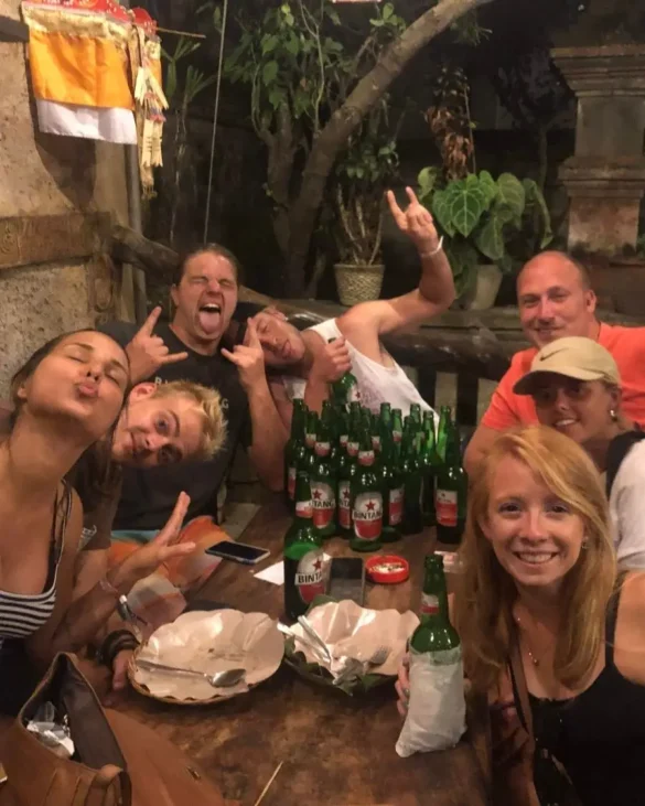 A group of 7 people with a large amount of beers at a table with plants in the back, posing and having a good time