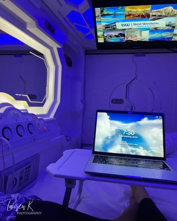 The inside of a pod bed with a TV on the ceiling, lighted mirror, purple lights to make it look futuristic and a bed tray with a laptop on it to show size