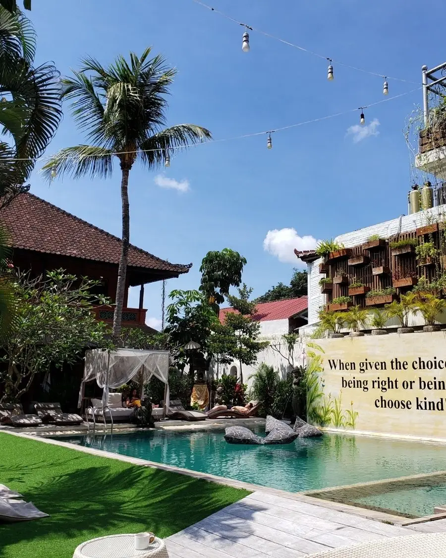 The pool deck with a giant mural on the wall of an inspirational quote, with string lights overhead and lots of plants under a sunny sky with people lounging