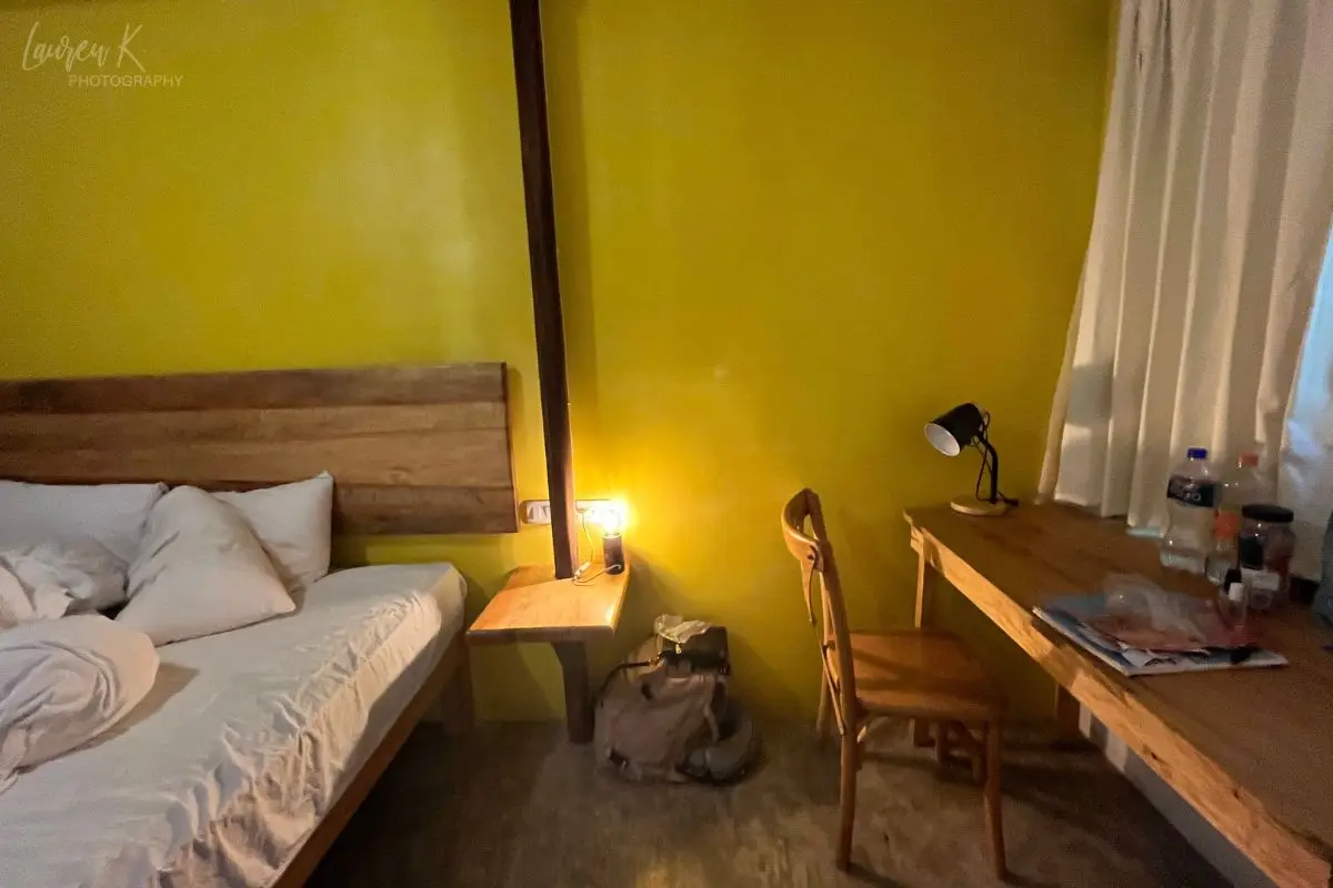 Part of a basic private room inside the Selina Puerto Escondido hostel with yellow wall, desk, nightstand and large bed