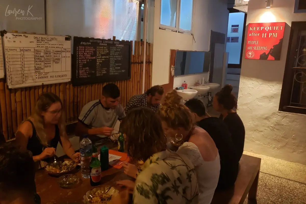 People eating dinner on a table with benches, in a dimly lit hostel common area, showing that checking hostels for free amenities is one of the best hostel tips to be aware of for travelers on a budget