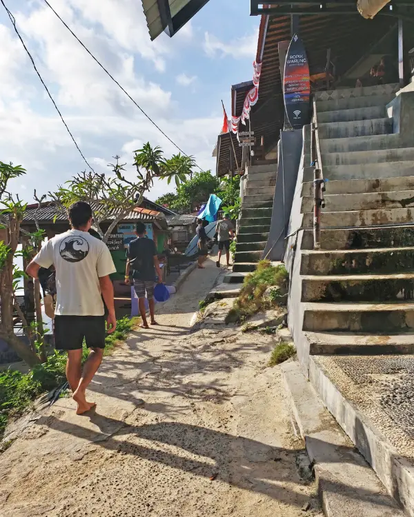 Man walking along the cliffside path at Suluban Beach, with warungs and shops on either side