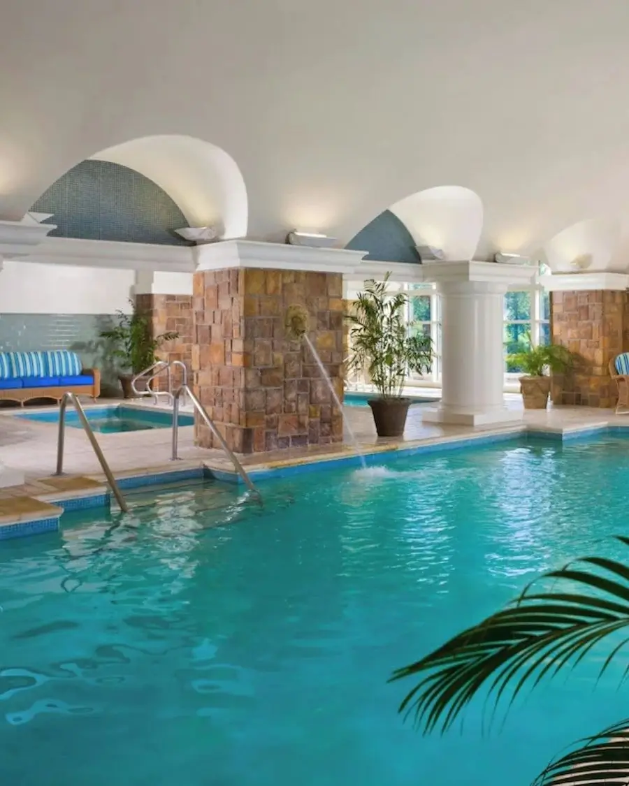 The indoor pool and hot tub area at the Ballantyne Resort with a palm tree off to the side and a jet shooting down from the stone wall