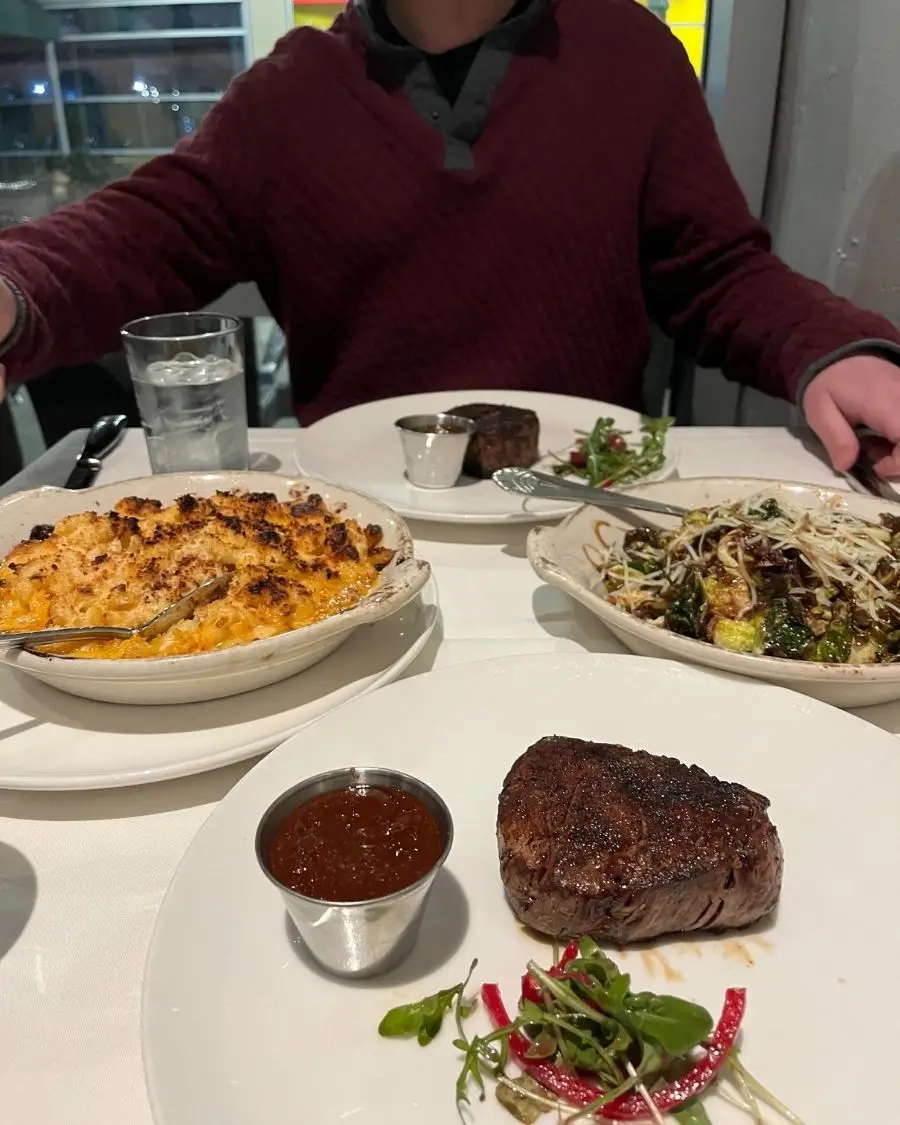 Juicy steaks, brussel sprouts and a big baked mac n cheese plate on a white tablecloth 