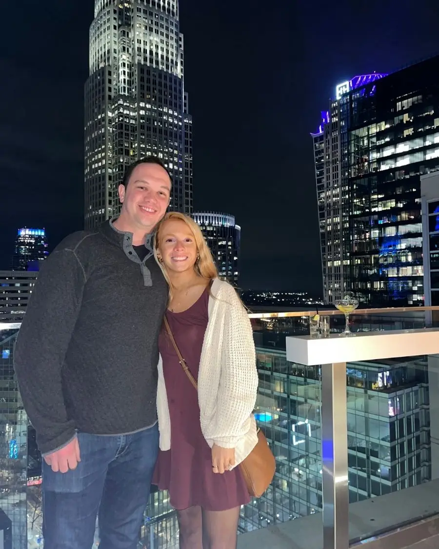 A couple smiling and posing in front of Charlotte high rises lit up at night, to show that Nuvole rooftop is one of the most romantic things to do in Charlotte