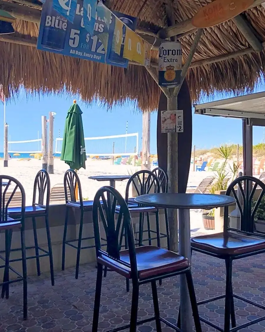 High top tables with chairs under a thatched roof with sand and the ocean in the background