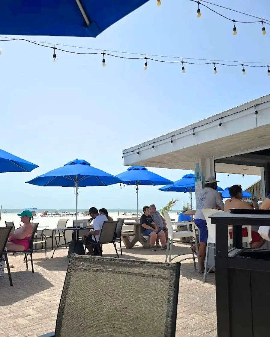 Umbrella tables in the patio of a bar under a sunny sky on the beach with string lights overhead and people relaxing