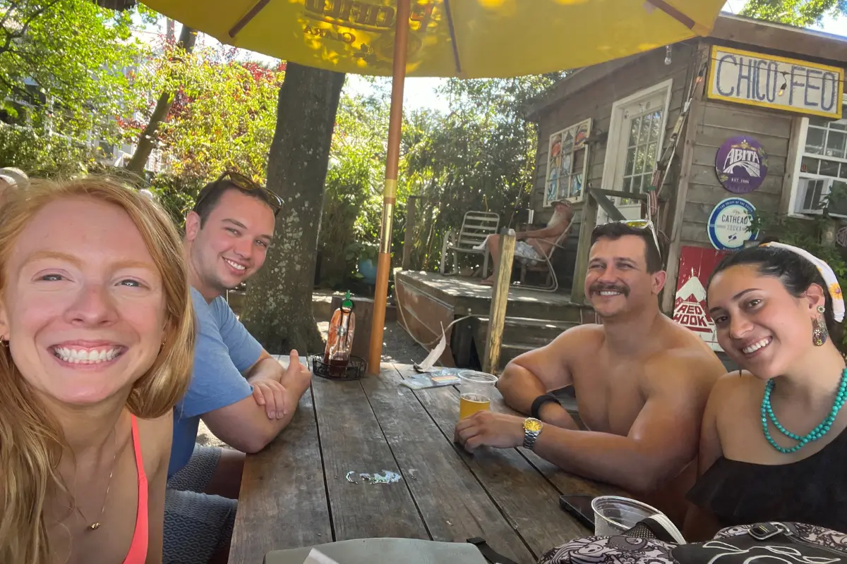 My husband and I, and a couple who are friends of ours all smiling, sitting in beachy clothes at a picnic table under the shade of trees at Chico Feo