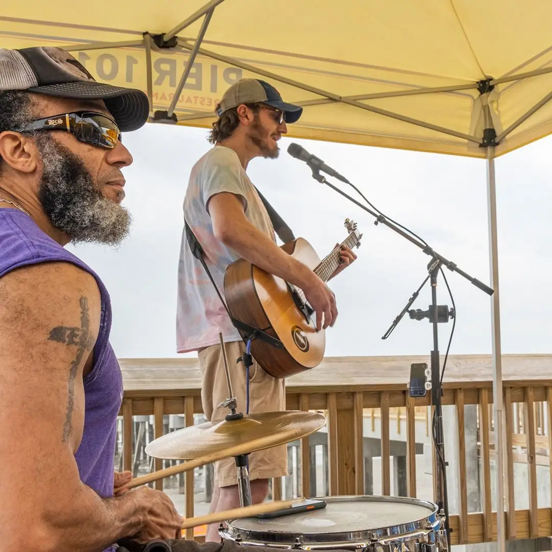 Man singing into a microphone with a guitar and a guy on drums next to him, underneath an umbrella at the Pier 101