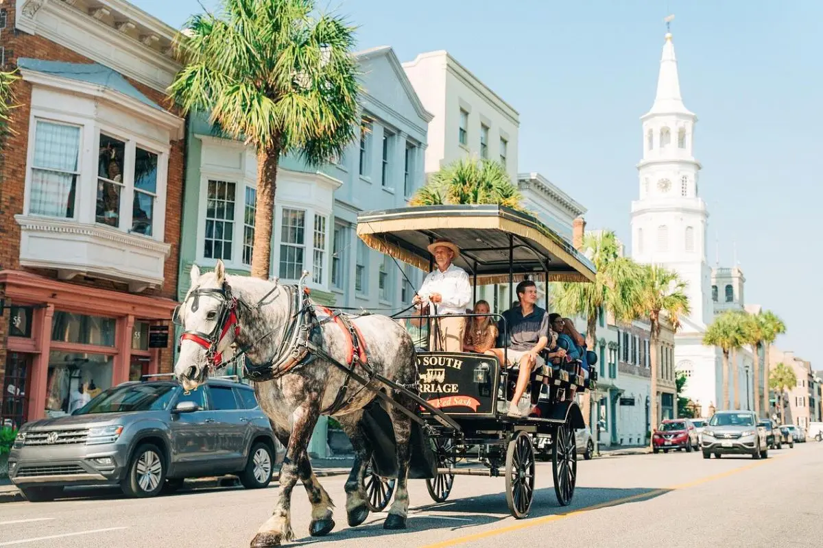 A horse-drawn carriage ride with people looking around, through the streets of Downtown Charleston on a sunny day