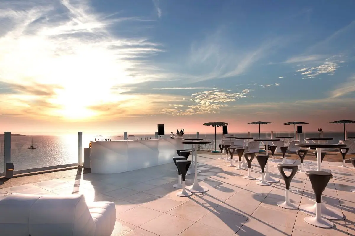 Modern, almost futuristic looking cocktail tables and a bar with marble flooring at a rooftop bar overlooking the sunset over the ocean