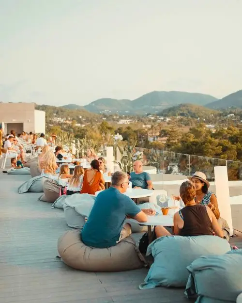 People lounging at tables sitting on bean bag style chairs overlooking the beautiful Ibiza island and hills, at the Rooftop Nine, which is one of the best Ibiza rooftop bars