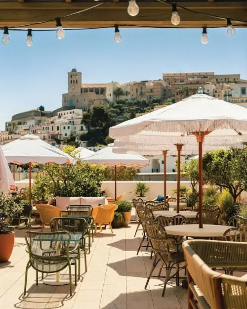 Tables with umbrellas at a bar overlooking Dalt Vila under string lights on a sunny day