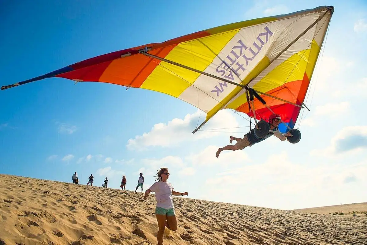 A person hang gliding with an instructor below over the dunes on a sunny day with a group of people looking on in the background