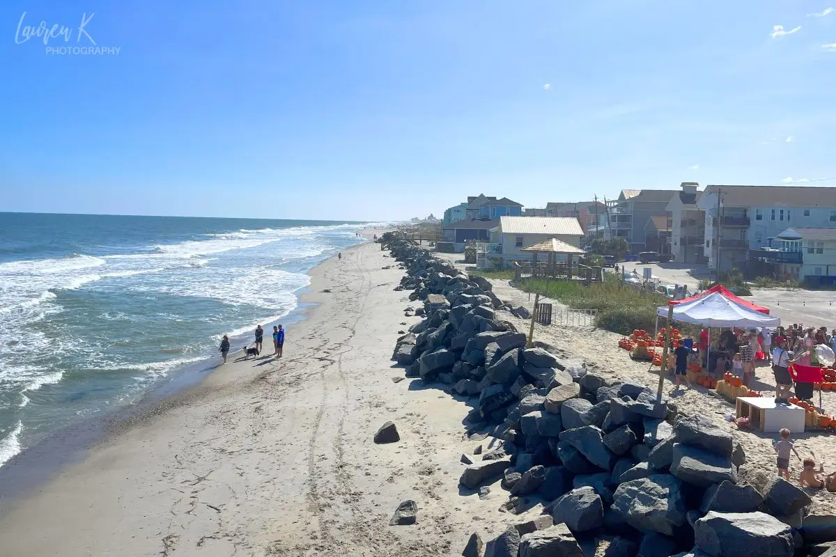 A view from the pier at Carolina Beach which shows the edge of the ocean, with rocks and then a fair going on above the beach on a sunny day