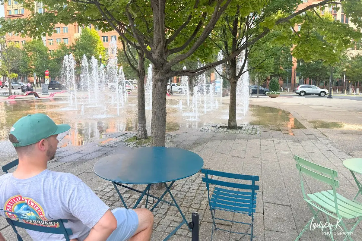 Cover image for the Washington DC Canal Park review by Inspired Backpacker, showing a man sitting at a patio table overlooking the park's fountains and trees