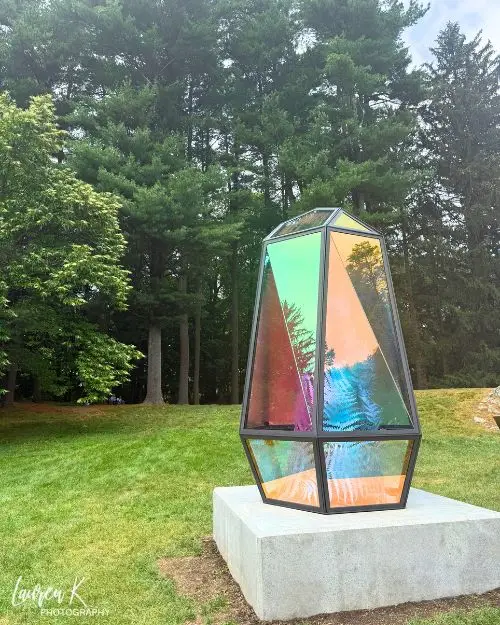 One of the pieces of art at the sculpture garden which is an iridescent prism, with woods behind and in a grassy green field