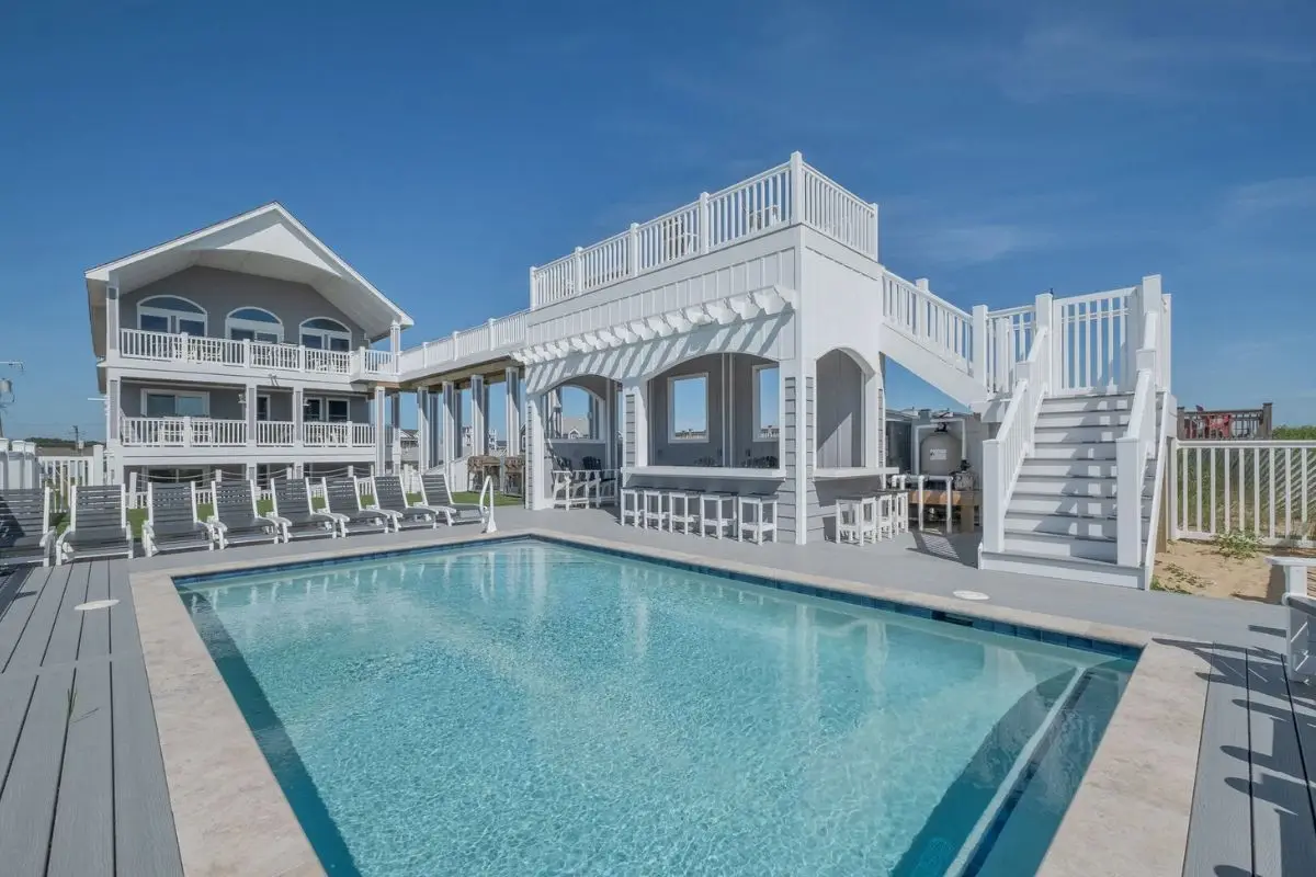 A huge rental house with a castle-like bar, a sky deck and sparkling pool on a sunny day, showing that a rental home is a great choice for large groups in terms of where to stay in Outer Banks NC