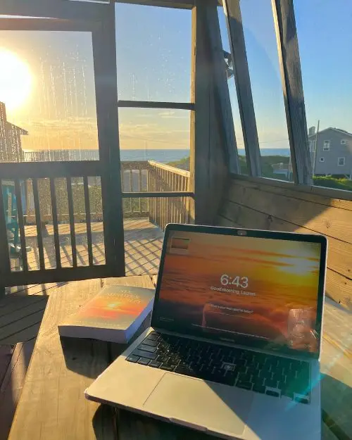 My computer and book on a picnic table inside a covered deck at our beach house, with the sunrise in the background