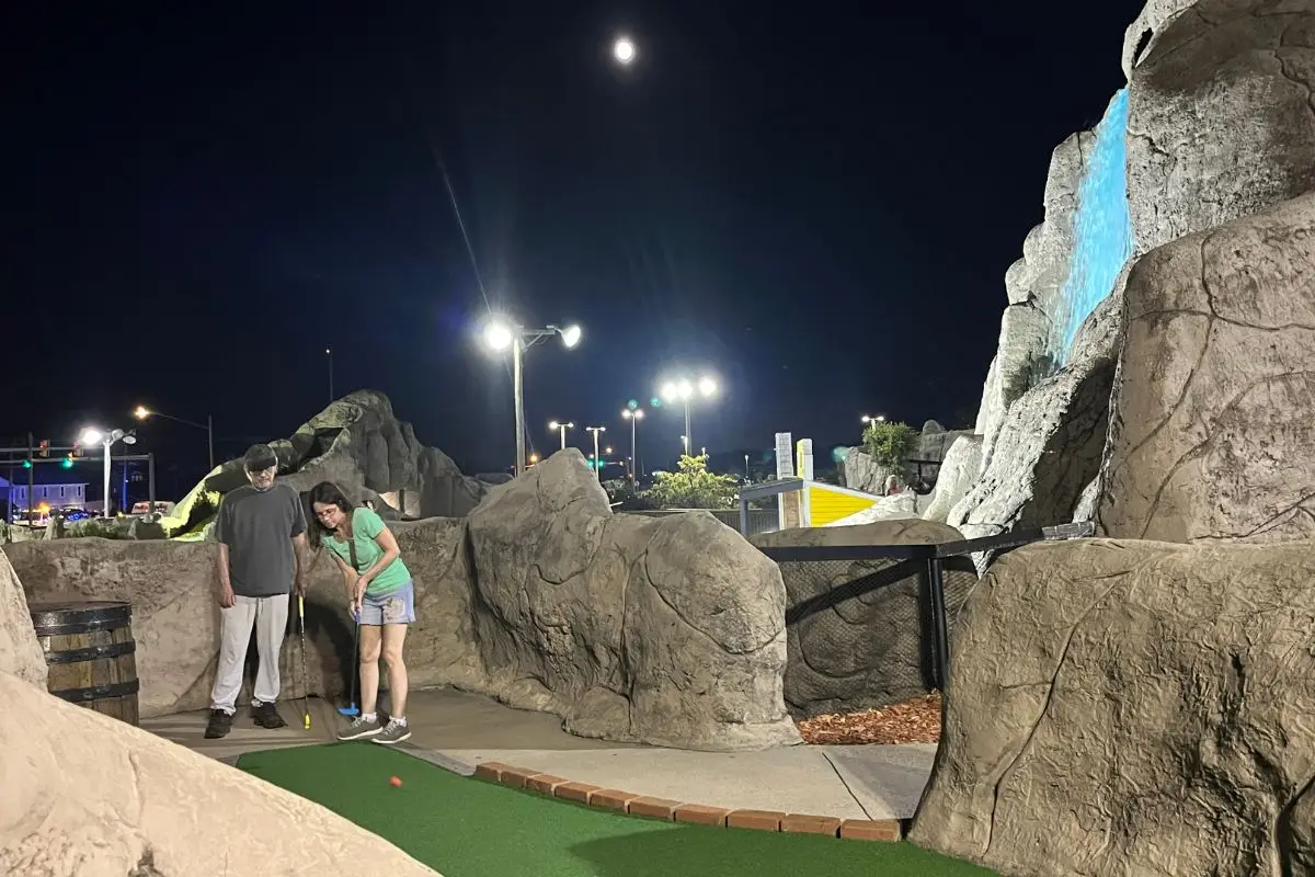 A couple about to hit their balls at a well-lit mini golf course under a full moon with a waterfall in the background