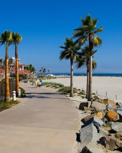 A curving boardwalk with a few people walking, palm trees blowing in the background and the beach at Coronado Beach island