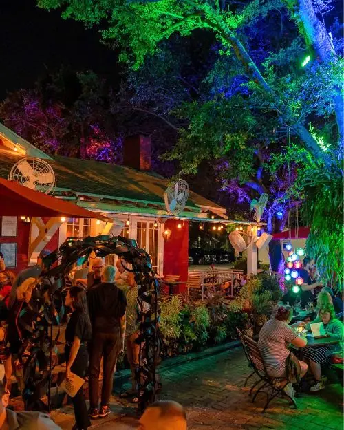 Trees lit up by neon lights overhead a patio and restaurant with string lights