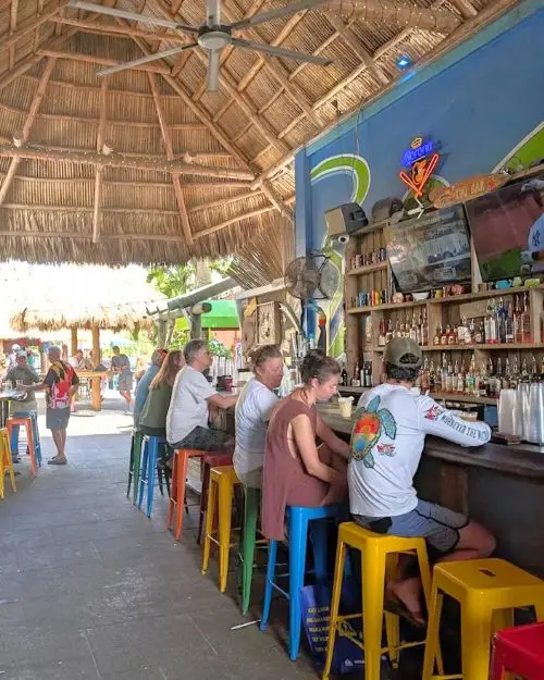 People sitting at a bar on colorful bar stools under a giant thatched roof during the day 
