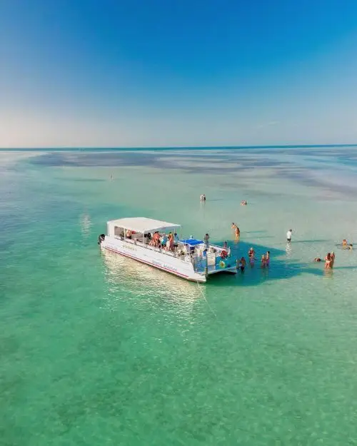 A boat in the middle of a teal, tropical looking ocean next to a sandbar with people out exploring on a cloudless sunny day