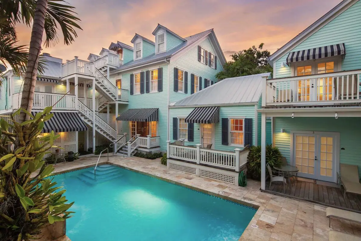 Two adjoined teal Victorian style homes underneath a sunsetting sky surrounded by palm trees and with the hotel pool in the courtyard, to show the general style of Key West and why it’s easily one of the best beach towns in Florida