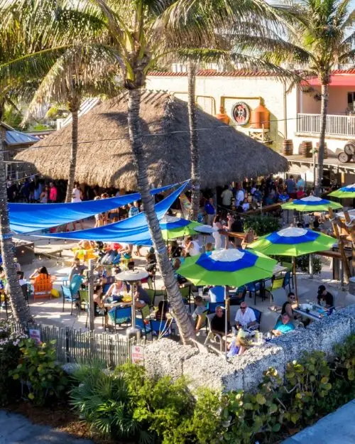 A crowded Sandbar, which is a bar in Delray Beach, with a big palapa, umbrellas and palm trees overhead, with people hanging out in the sand