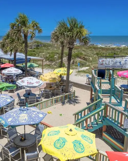An oceanfront restaurant and bar with colorful umbrella shaded tables and a playground with palm trees, with the ocean in the background