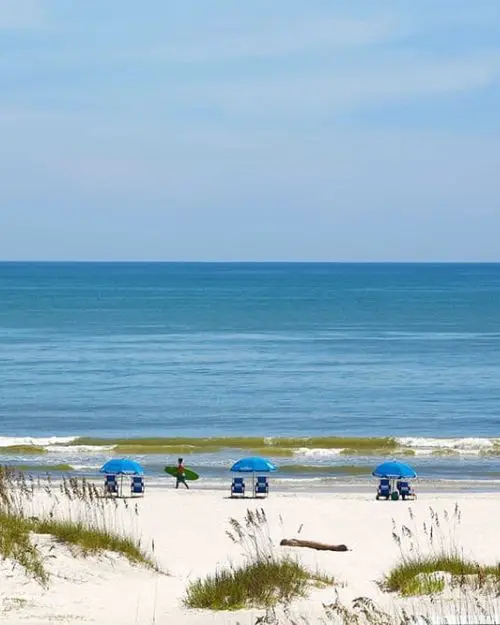 Surfer walking down the beach with beach umbrellas and chairs, with a wavy ocean in the background under a sunny sky
