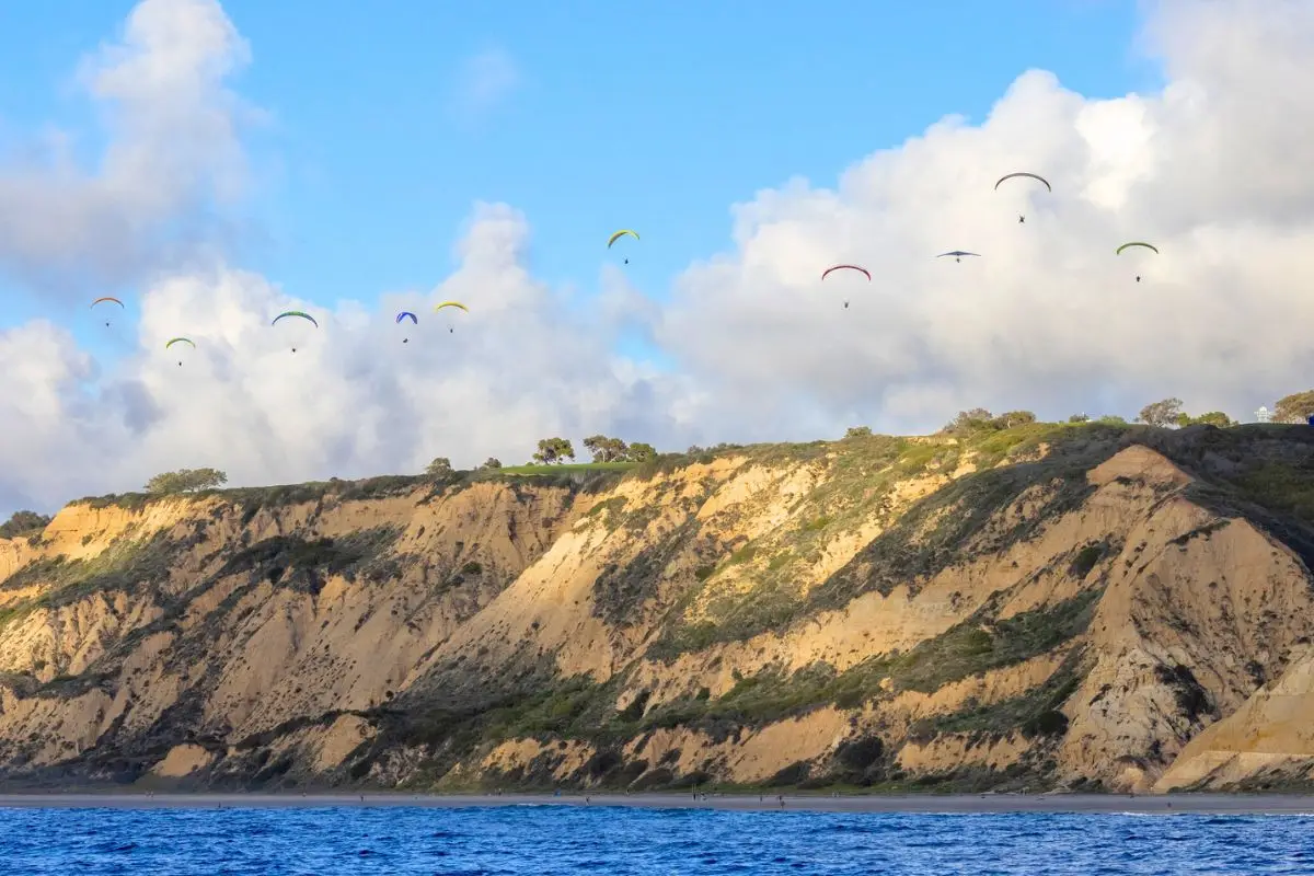 Gorgeous cliffs over the ocean with sun shining through some clouds, causing beautiful shadows along the cliffs, and a bunch of hang gliders above the cliffs