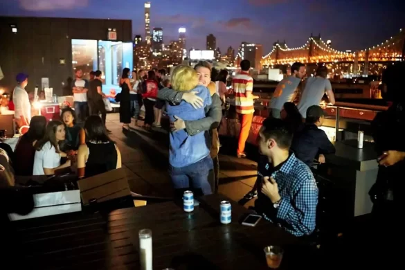 The buzzing rooftop of a hostel in NYC with lots of people casually enjoying themselves under the night time glow of NYC