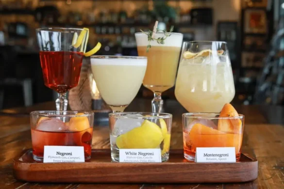Four beautiful cocktails presented on a tray with three glasses of fruit and juice, which is a flight offering by Artisan's Palate