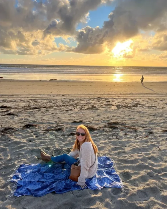 A redheaded woman on the beach during the sunset with pants, a sweater and on a blanket