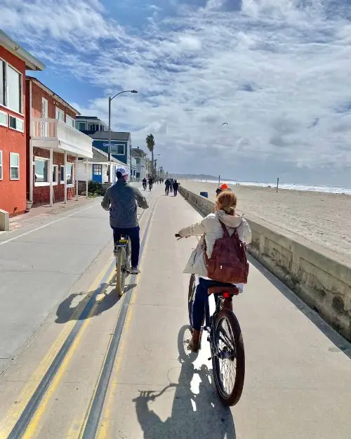 A couple biking down the Pacific Beach boardwalk on a sunny day with jackets