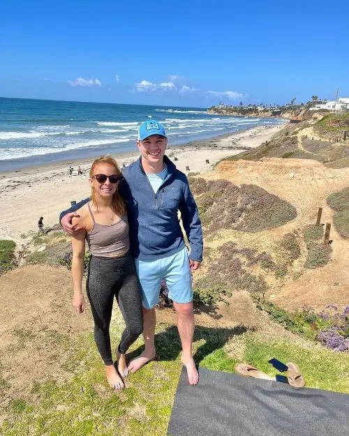 A couple smiling at the camera with athletic outfits on and the ocean in the background, on a cliff with their yoga mat after a yoga class on a sunny day