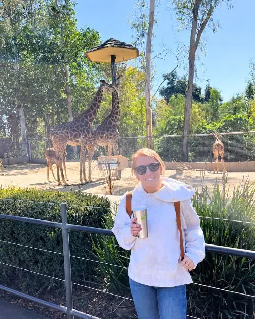 A redheaded woman with a drink in hand in front of some giraffes on a sunny day at the San Diego zoo