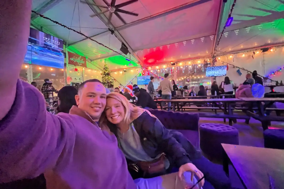 A happy couple taking a selfie in a tented bar on a couch with glowing lights behind them and communal style tables