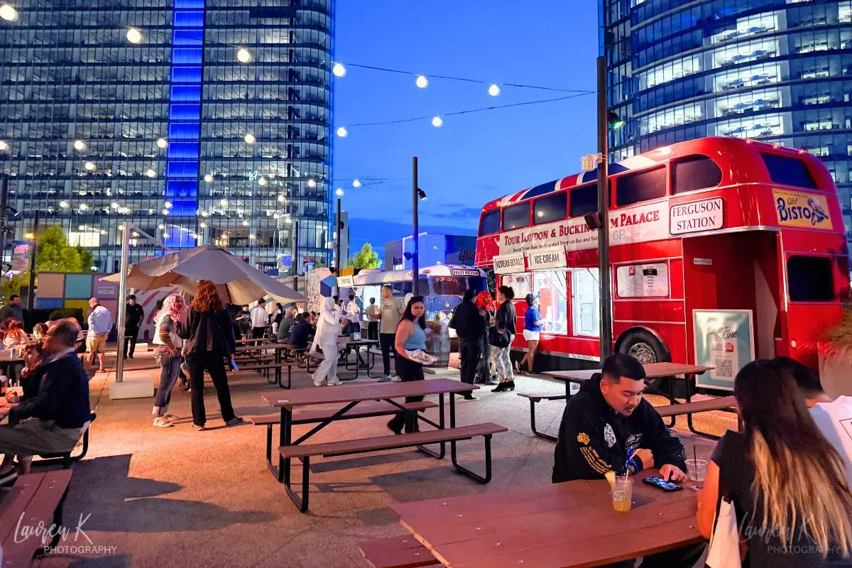Food trucks including one which is shaped like a double decker bus, underneath string lights and high rise buildings