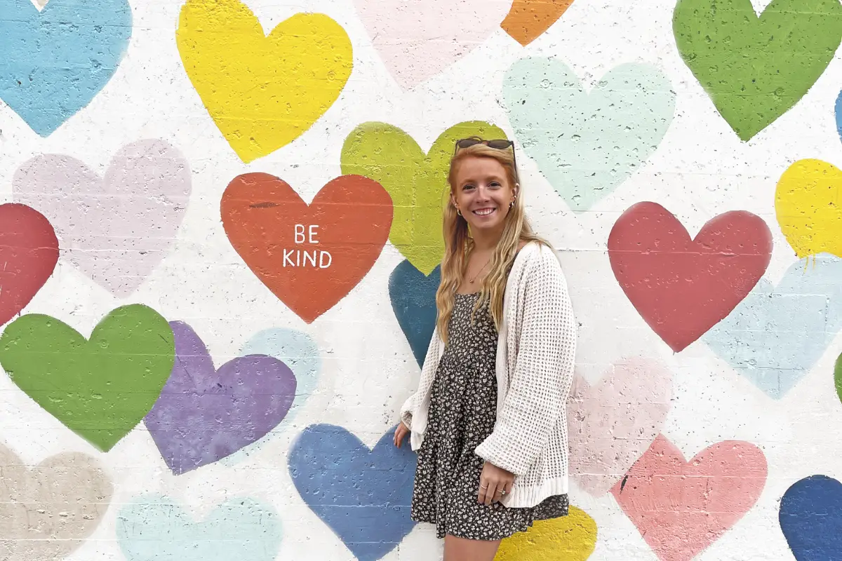 The Confetti Hearts mural wall in NoDa Charlotte with the wording Be Kind, with Inspired Backpacker Lauren Kessel standing in front with a smile
