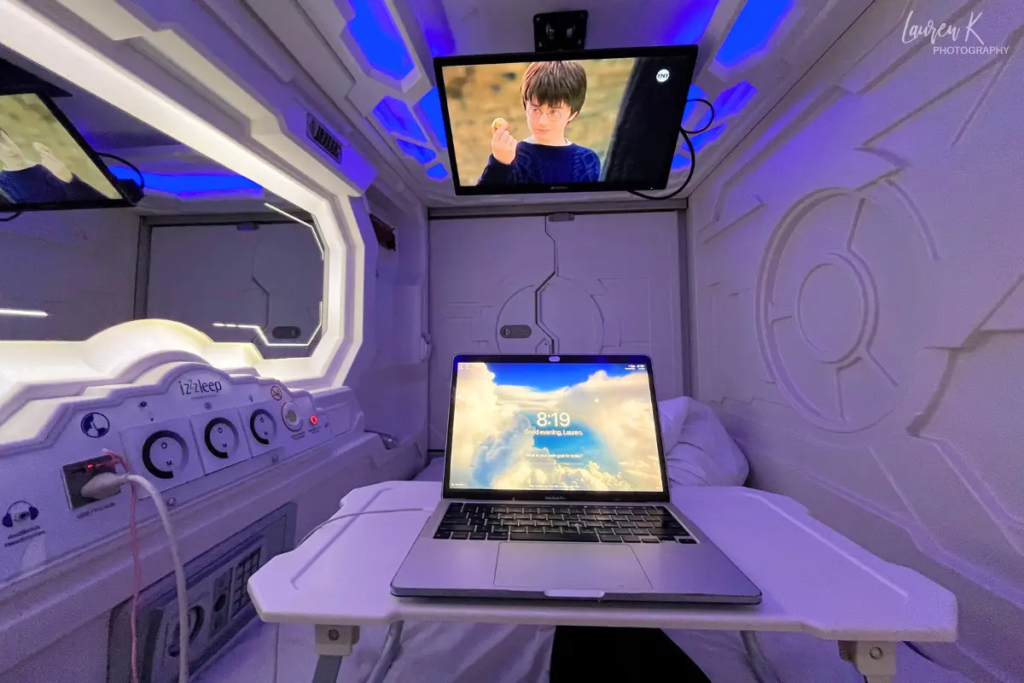 POV photo at the IzZzleep Aeropuerto Terminal 1 hotel at the Mexico City Airport, glowing in purple light with a laptop out and TV playing above, very futuristic looking